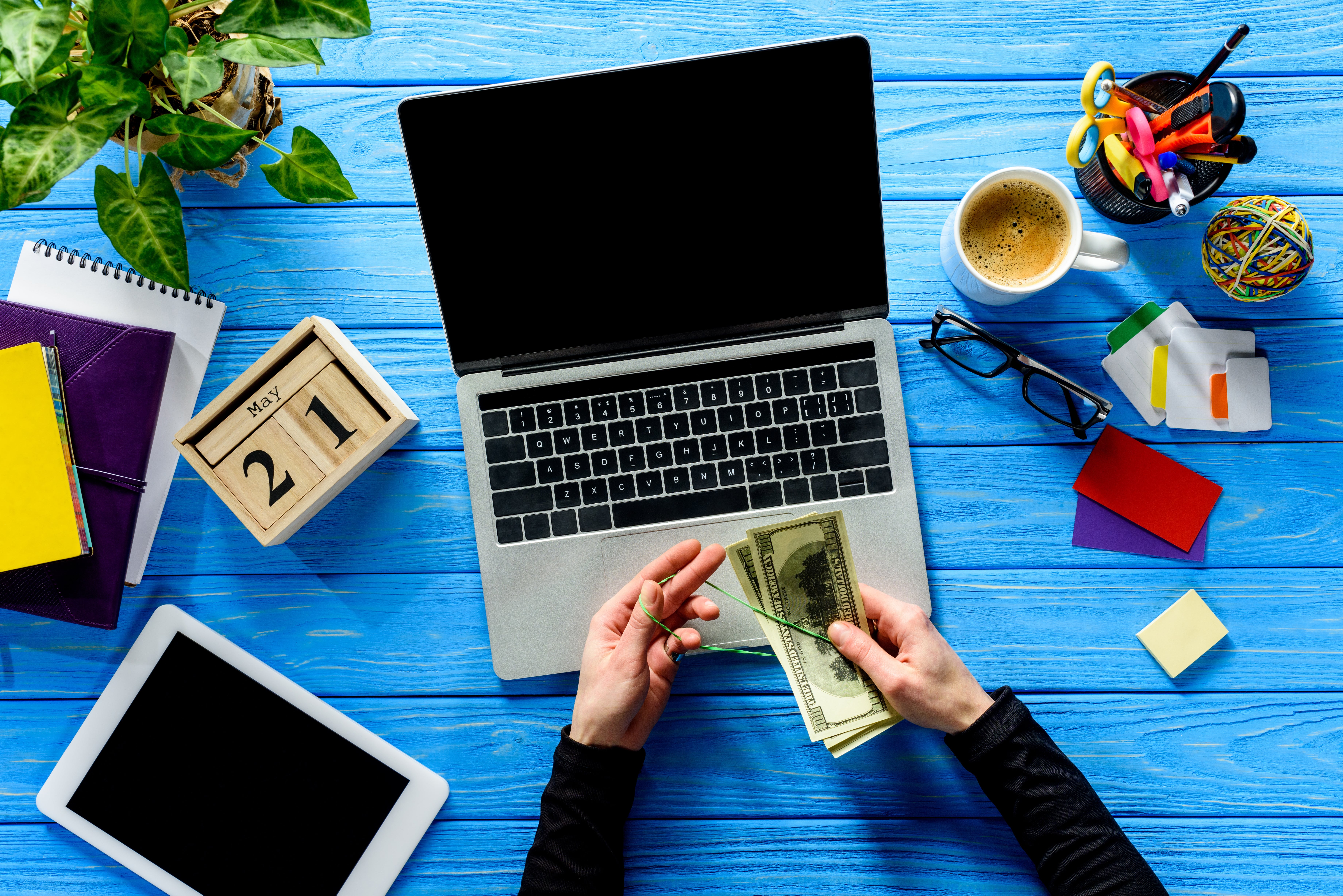 a hand counting money with a laptop, ipad, and coffee in the background