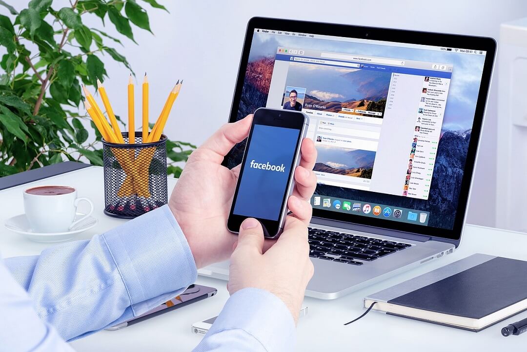 Man in front of a laptop holding his phone checking Facebook