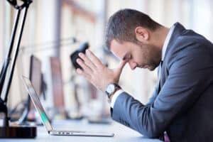 frustrated young businessman sitting at a desk behind a laptop with his hands up to his head